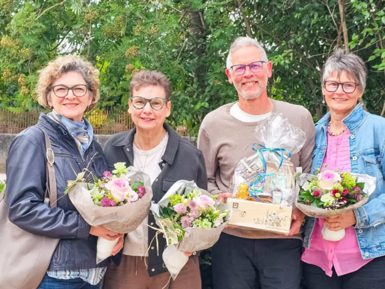 Drei Frauen und ein Mann stehen lächelnd nebeneinander vor Sträuchern, sie halten Blumensträusse und ein Geschenkkorb.