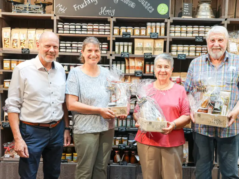Vier lächelnde Personen stehen mit Geschenkkörben in einem Laden vor Regalen mit Lebensmitteln.