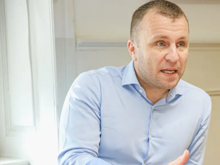 Ein Mann im hellblauen Hemd sitzt an einem Tisch und spricht mit gestikulierenden Händen.