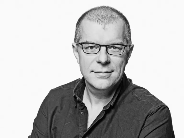 Schwarz-weiß-Porträt eines kurzhaarigen Mannes mit Brille und dunklem Hemd, der in die Kamera blickt.