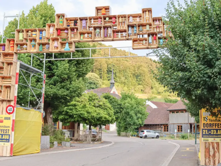Ein Torbogen aus bunt bemalten Holzkisten überspannt eine Dorfstrasse vor grünen Hügeln.