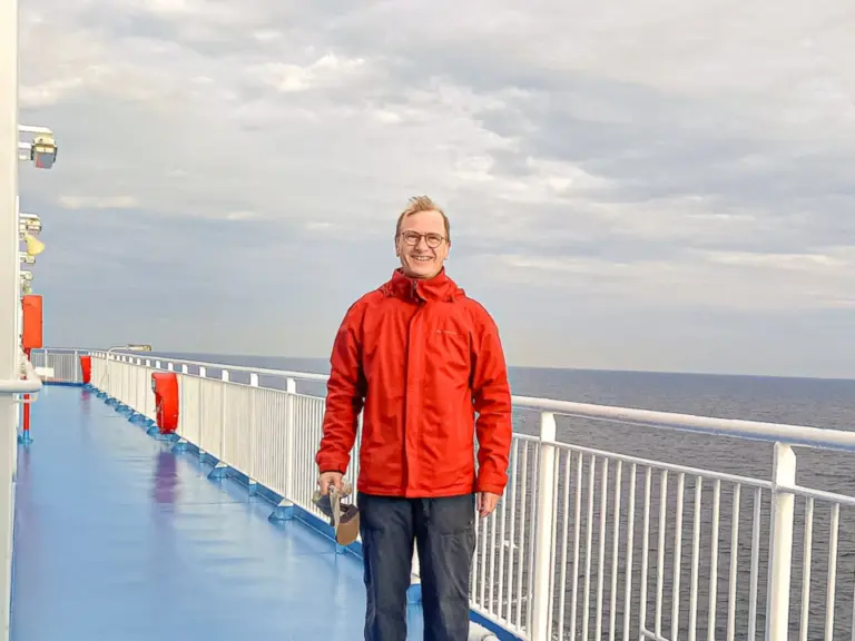Ein Mann in roter Jacke steht auf dem blauen Deck eines Fährschiffs vor dem offenen Meer.