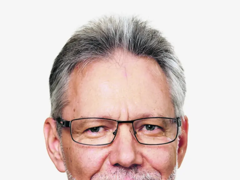 Ein älterer Mann mit grauem Haar, Brille und Bart trägt Anzug mit Krawatte und lächelt.