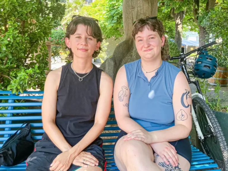 Zwei lächelnde Frauen sitzen nebeneinander auf einer blauen Bank unter einem Baum, daneben ein Fahrrad.
