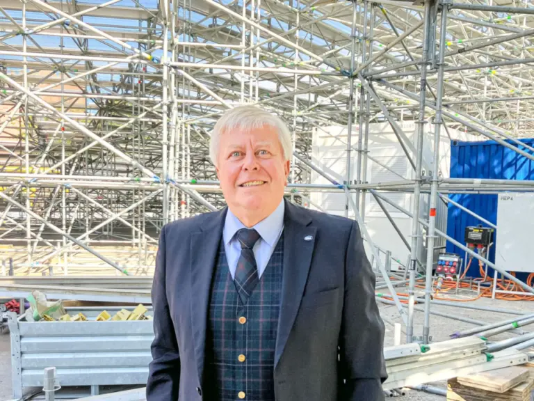Ein lächelnder Mann im Anzug mit Karo-Weste steht vor einer Baustelle mit Gerüsten.
