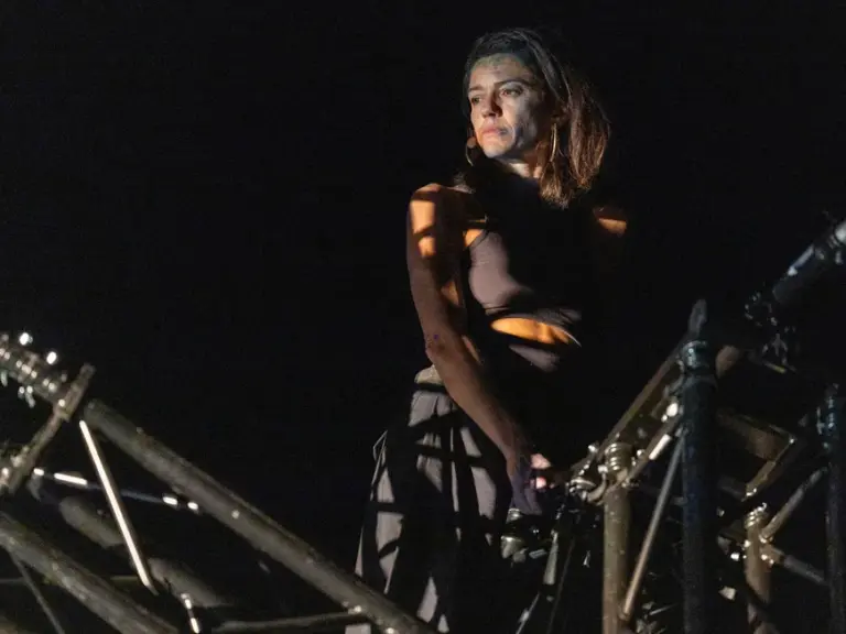 Eine Frau steht nachts im Scheinwerferlicht zwischen Metallgerüsten auf einer Bühne.