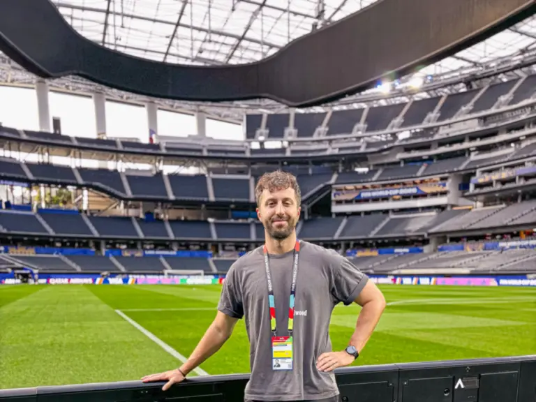 Ein Mann mit Akkreditierung steht lächelnd am Spielfeldrand eines leeren Fussballstadions.