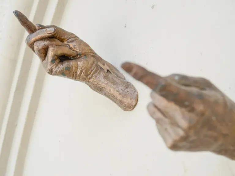 Zwei bronzene Hände strecken sich mit ausgestrecktem Zeigefinger einander entgegen, ohne sich zu berühren.