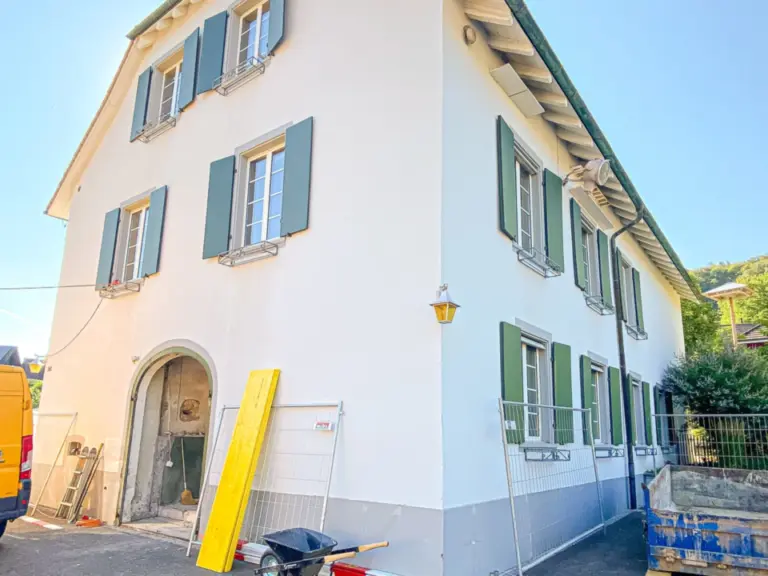 Weisses Haus mit grünen Fensterläden während Renovierungsarbeiten, mit Baugerüst und Bauzaun davor.
