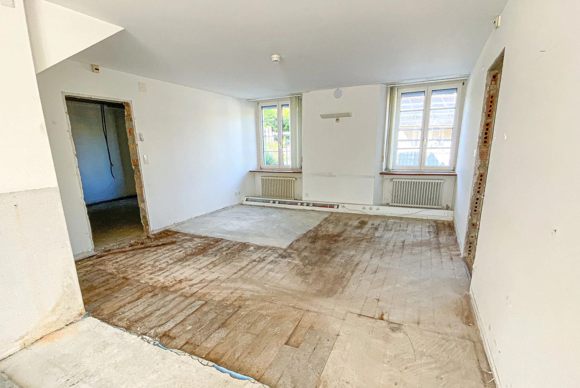 Leerer Raum mit freigelegtem Dielenboden, weissen Wänden und zwei Sprossenfenstern während einer Renovierung.