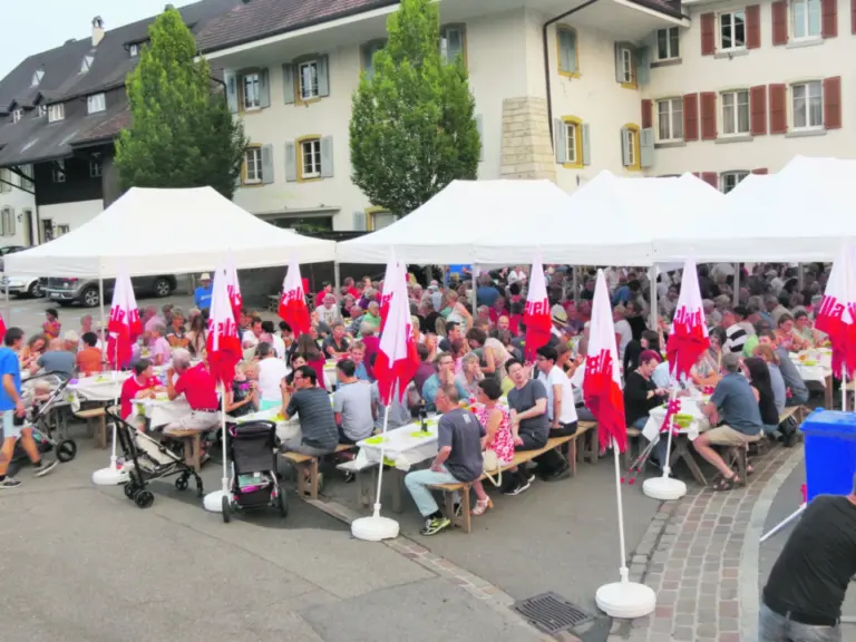 Menschen sitzen an langen Tischen unter weissen Zelten mit roten Fahnen bei einem Dorffest.