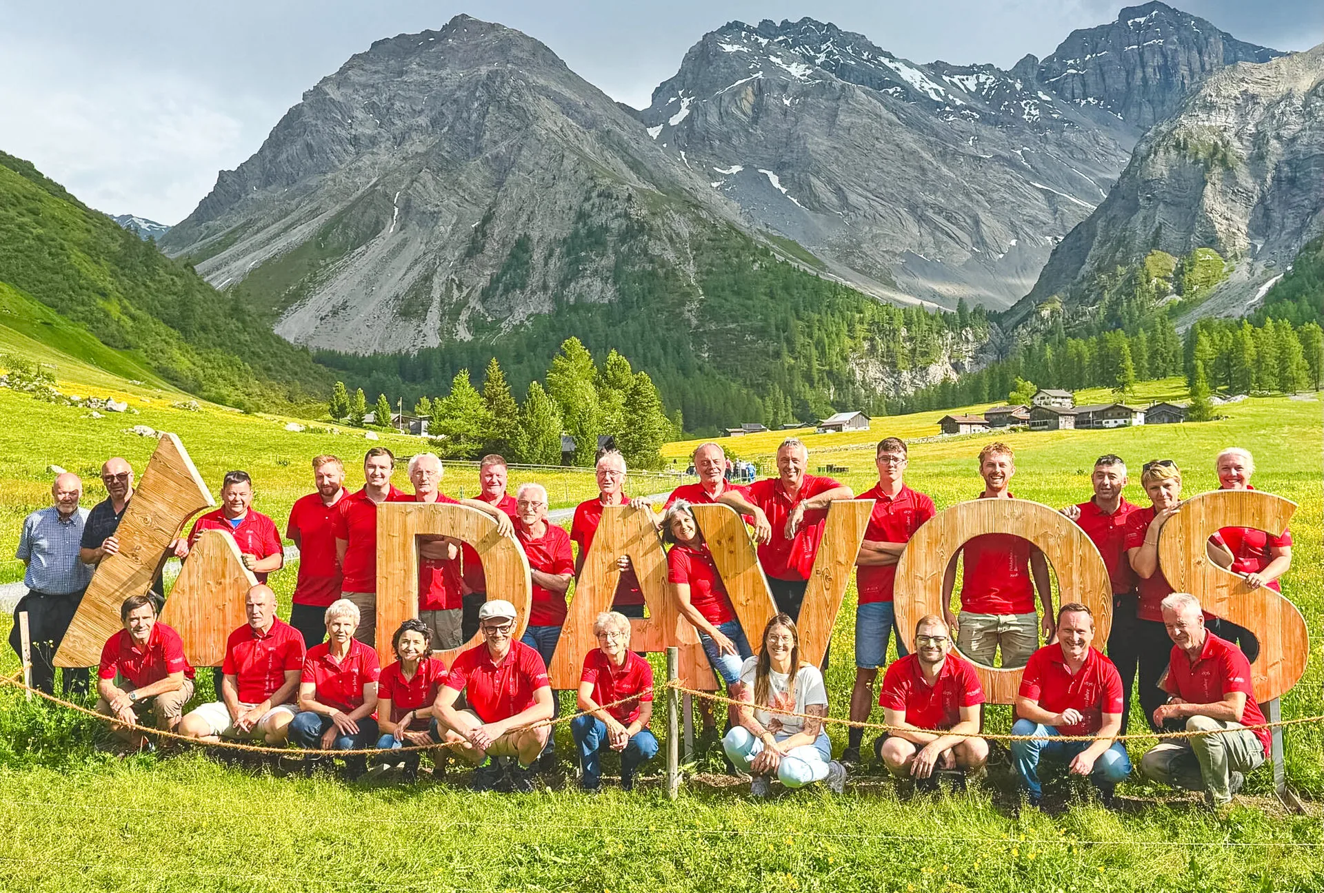 Grosse Gruppe in roten Poloshirts posiert um hölzerne Buchstaben "DAVOS" in einem grünen Alpental vor Bergpanorama.