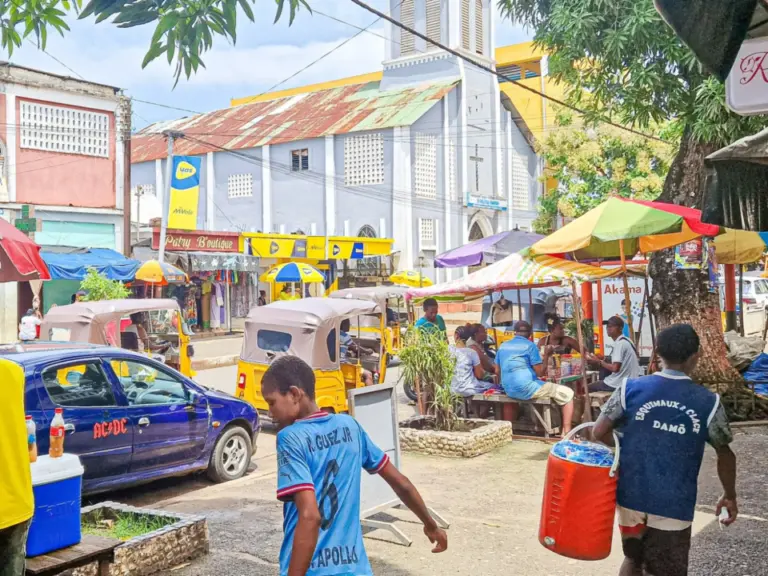 Zwei Männer gehen über eine Marktstrasse, im Hintergrund Stände, Tuk-Tuks und eine weisse Kirche mit Turm.