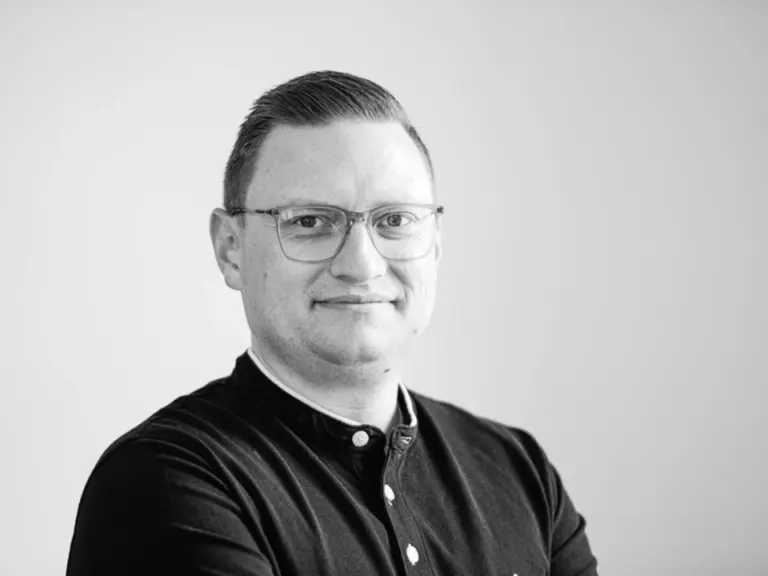 Schwarz-weiß-Porträt eines lächelnden Mannes mit Brille in dunklem Hemd, verschränkten Armen, vor hellem Hintergrund.