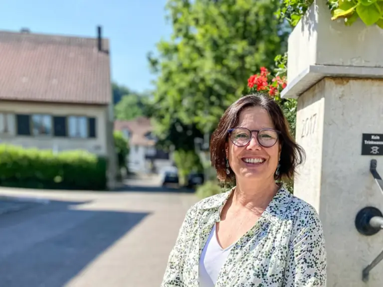 Lächelnde Frau mittleren Alters sitzt an einem steinernen Dorfbrunnen mit Blumen in der Sonne.