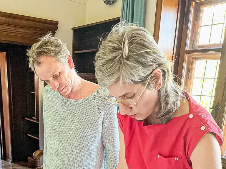 Ein Mann und eine Frau beugen sich über einen Tisch mit Karten und Zeichnungen in einem Zimmer mit Holzvertäfelung.