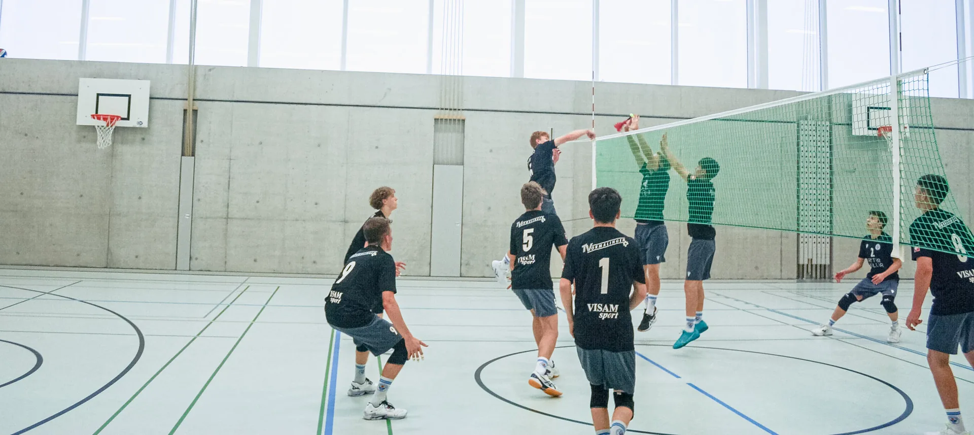 Jugendliche spielen Volleyball in einer Sporthalle, ein Spieler springt zum Schmetterschlag am Netz.