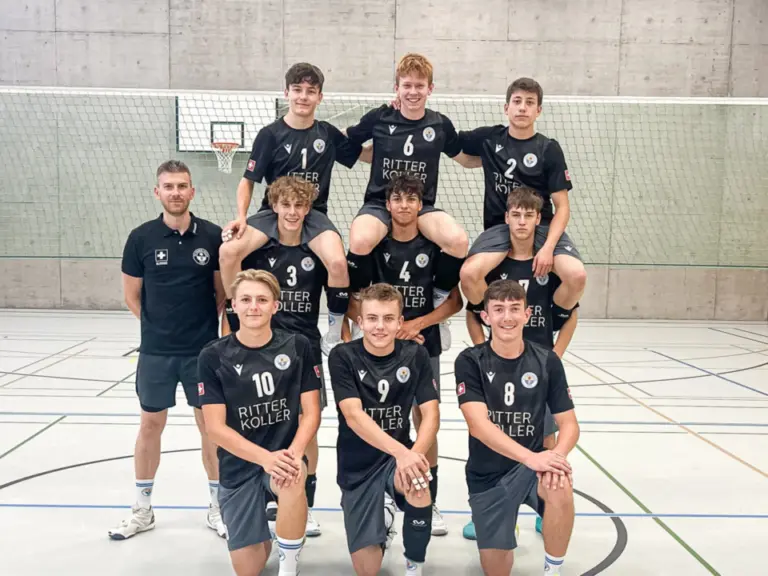 Eine Jugend-Volleyballmannschaft in schwarzen Trikots posiert in drei Reihen in einer Sporthalle.
