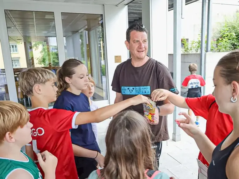 Ein Mann steht mit mehreren Kindern im Kreis vor einem Gebäudeeingang und verteilt kleine Gegenstände.