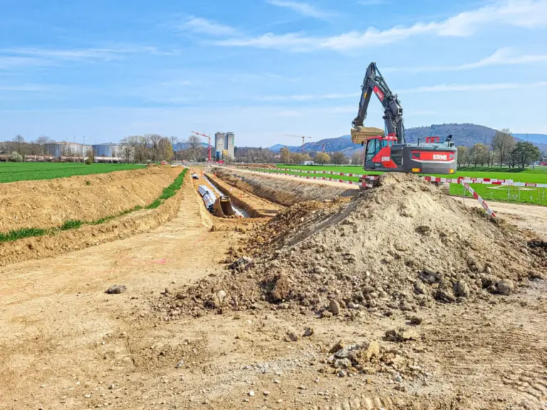 Bagger gräbt einen langen Erdgraben mit verlegtem Rohr neben einem Feld, Hügel im Hintergrund.