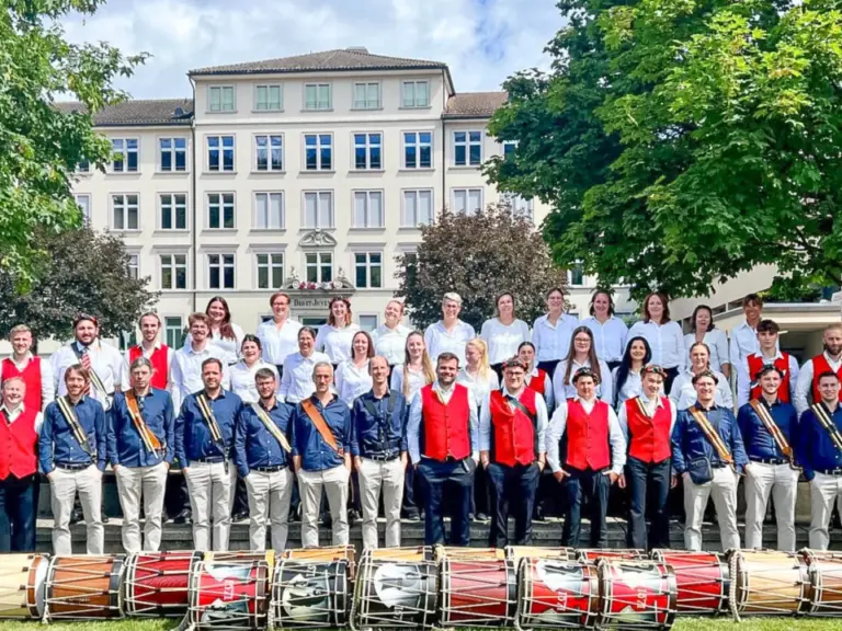 Gruppenfoto einer Trommler- und Chorformation in Trachten vor einem Gebäude, mit Trommeln auf dem Rasen davor.