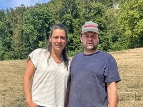 Eine Frau und ein Mann stehen nebeneinander auf einer trockenen Wiese vor bewaldeten Hügeln.