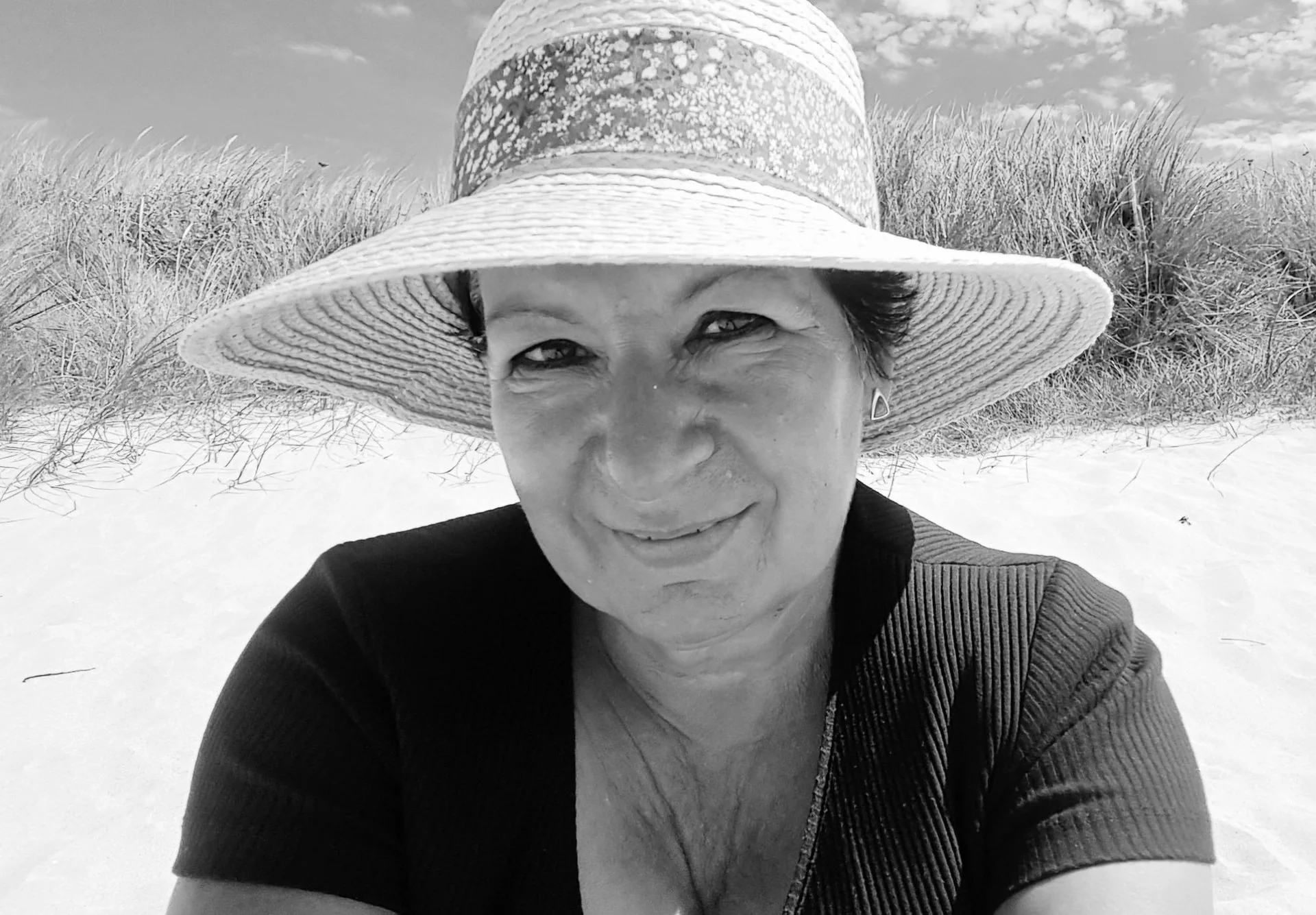 Lächelnde Frau mit Sonnenhut sitzt an einem Sanddünenstrand, Schwarzweissfoto.