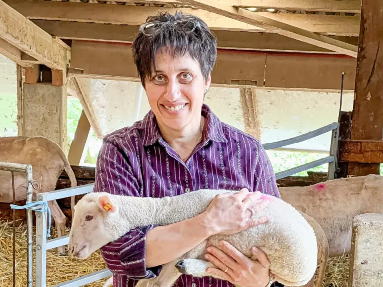 Eine lächelnde Frau hält ein Lamm im Arm, umgeben von weiteren Schafen in einem Stall mit Stroh.