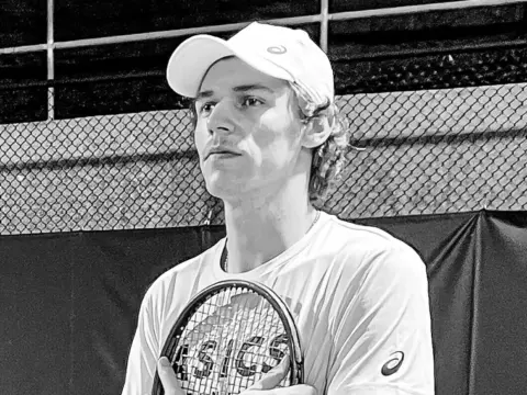 Ein junger Mann mit weisser Kappe hält einen Tennisschläger vor der Brust und blickt zur Seite.