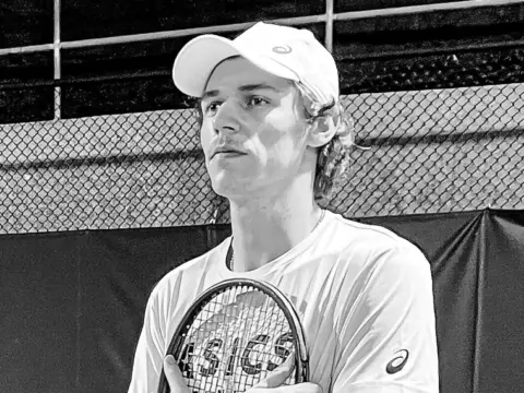 Ein junger Tennisspieler mit Cap hält einen Schläger an die Brust und blickt zur Seite, Schwarzweißfoto.