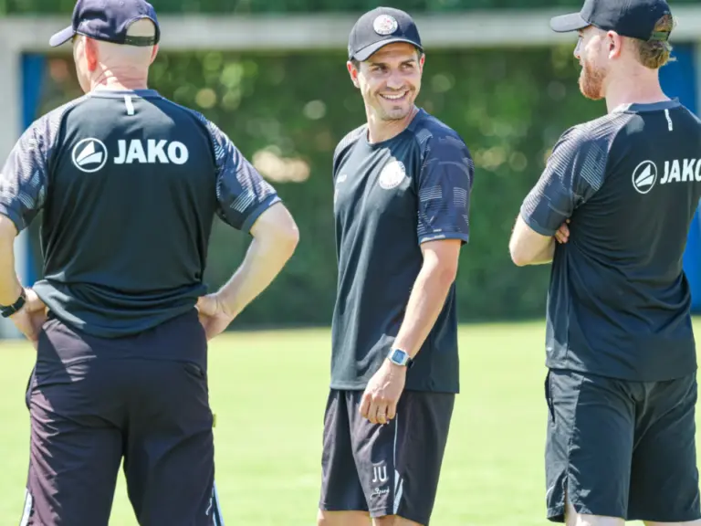 Drei Trainer in dunklen Trikots mit Cap stehen lachend auf einem Fussballplatz zusammen.