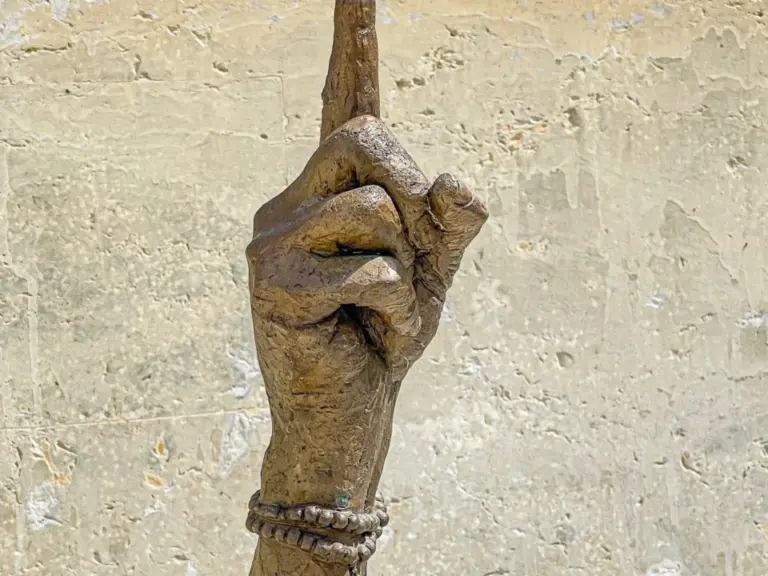 Eine bronzene Skulptur einer erhobenen Hand mit Armreif, auf deren Zeigefinger eine kleine Figur balanciert.