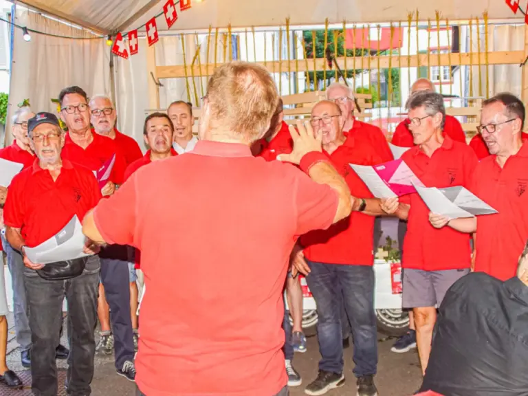 Männerchor in roten Shirts singt unter einem Zeltdach, ein Dirigent in Rot leitet vor ihnen.
