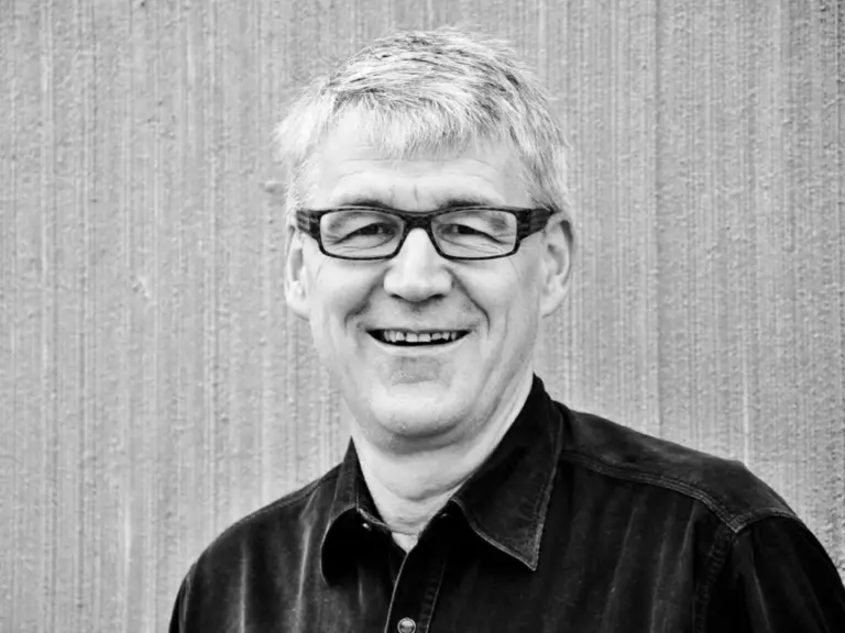 Ein lächelnder älterer Mann mit grauem Haar und Brille, in dunklem Hemd, vor einer strukturierten Wand.