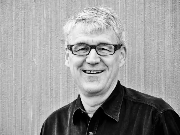 Ein lächelnder älterer Mann mit Brille und grauem Haar in dunklem Hemd vor gestreifter Wand.