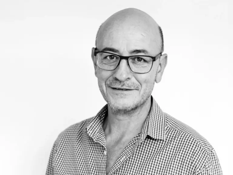 Ein älterer Mann mit Brille, Halbglatze und kariertem Hemd blickt lächelnd in die Kamera.