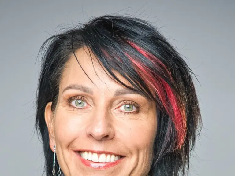Lächelnde Frau mit schwarzen, rot gesträhnten Haaren in weissem Rollkragenpullover und dunklem Blazer.