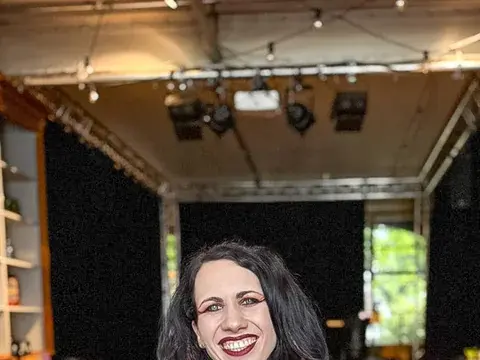 Eine lachende Frau mit schwarzen Haaren und roten Dreadlock-Strähnen sitzt an einem Tisch in einem Veranstaltungsraum.