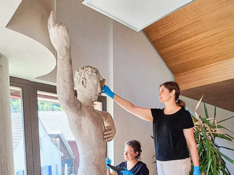 Zwei Frauen mit blauen Handschuhen reinigen eine lebensgrosse Steinstatue eines nackten Mannes in einem Raum.