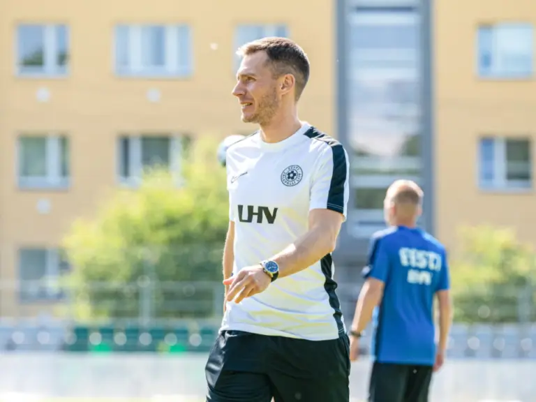 Ein Mann im Trainingsshirt lächelt und blickt zur Seite auf einem Sportplatz, im Hintergrund ein weiterer Mann.