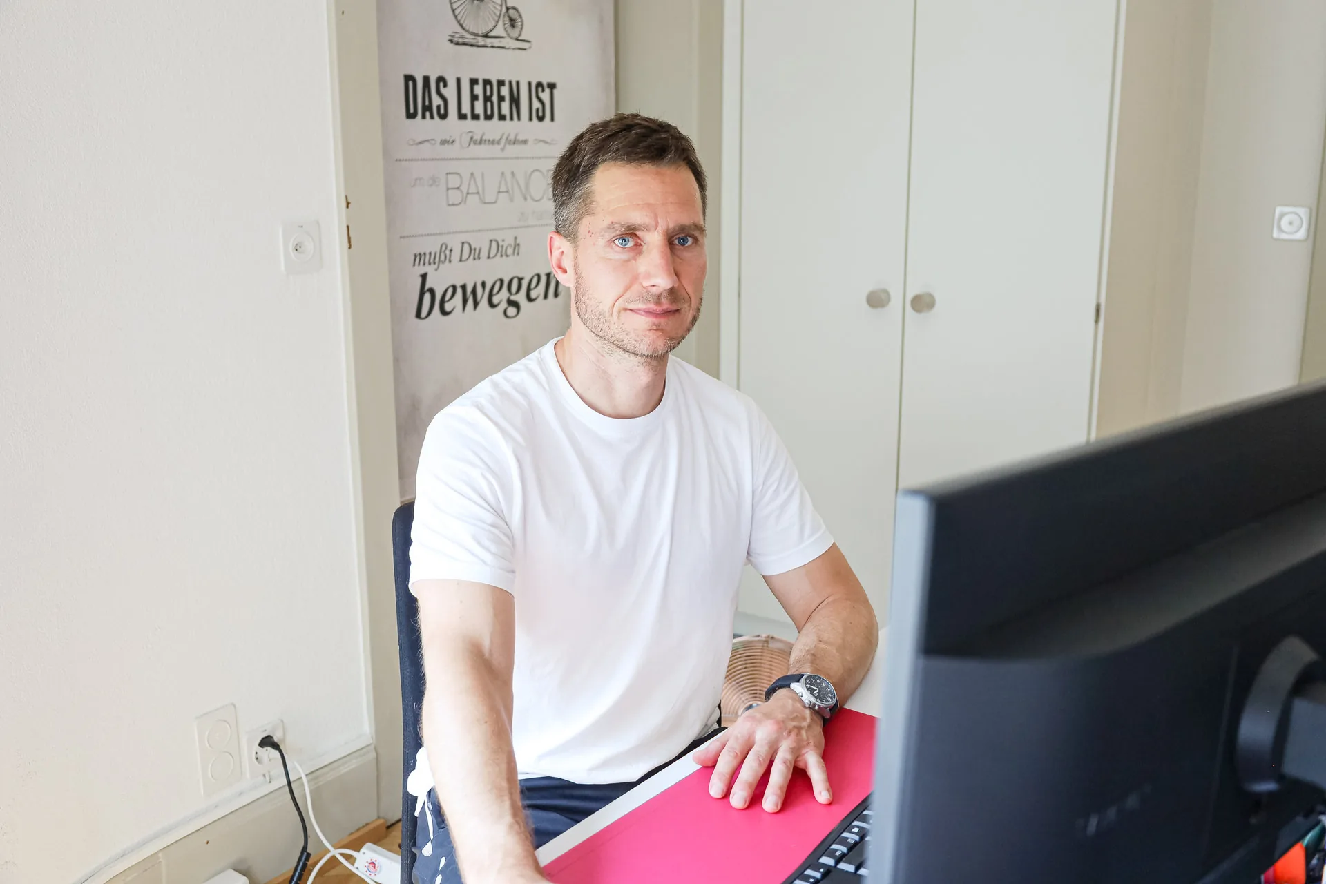 Ein Mann im weissen T-Shirt sitzt an einem Schreibtisch vor einem Computerbildschirm und blickt in die Kamera.