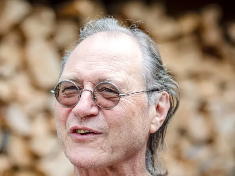Ein älterer Mann mit runder Brille und grauem Haar lächelt vor einem Holzstapel im Hintergrund.