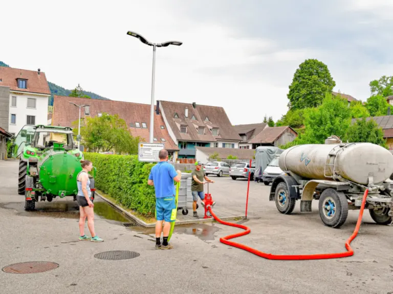 Personen befüllen einen Tankwagen mit rotem Schlauch neben grünen Traktoren auf einem Dorfplatz.