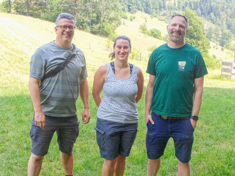 Drei lächelnde Erwachsene stehen nebeneinander auf einer grünen Wiese vor bewaldeten Hügeln.
