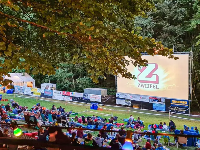 Zuschauer sitzen auf Decken im Freien vor einer Leinwand mit Werbebanner unter Bäumen bei Dämmerung.