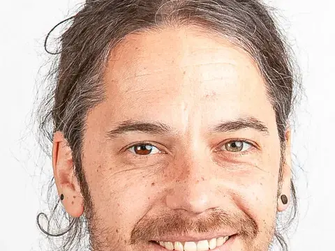 Ein lächelnder Mann mit zusammengebundenen Haaren und Bart trägt ein geblümtes Hemd.