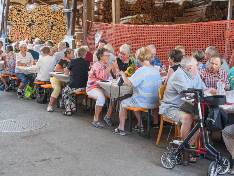 Ältere Menschen sitzen an langen Tischen im Freien unter einem Holzschuppen und unterhalten sich beim Essen.