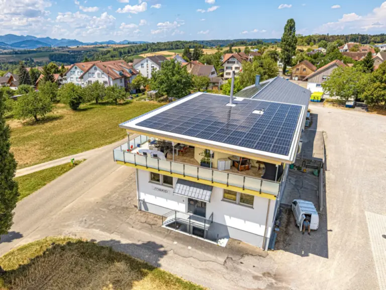 Luftaufnahme eines Gebäudes mit grossflächiger Photovoltaikanlage auf dem Dach, umgeben von grüner Landschaft.