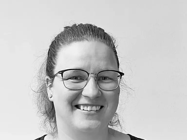 Eine lächelnde Frau mittleren Alters mit Brille und zurückgebundenem Haar in Schwarz-Weiß.