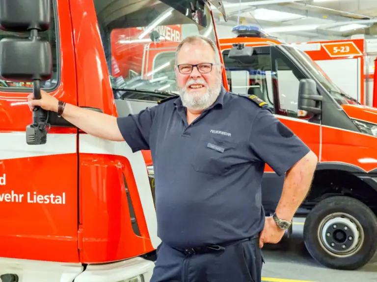 Ein lächelnder Mann mit Brille und Bart steht an ein Feuerwehrfahrzeug gelehnt in einer Fahrzeughalle.